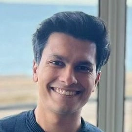 Smiling man with dark hair in a blue sweater, standing by a window with a blurred ocean view.